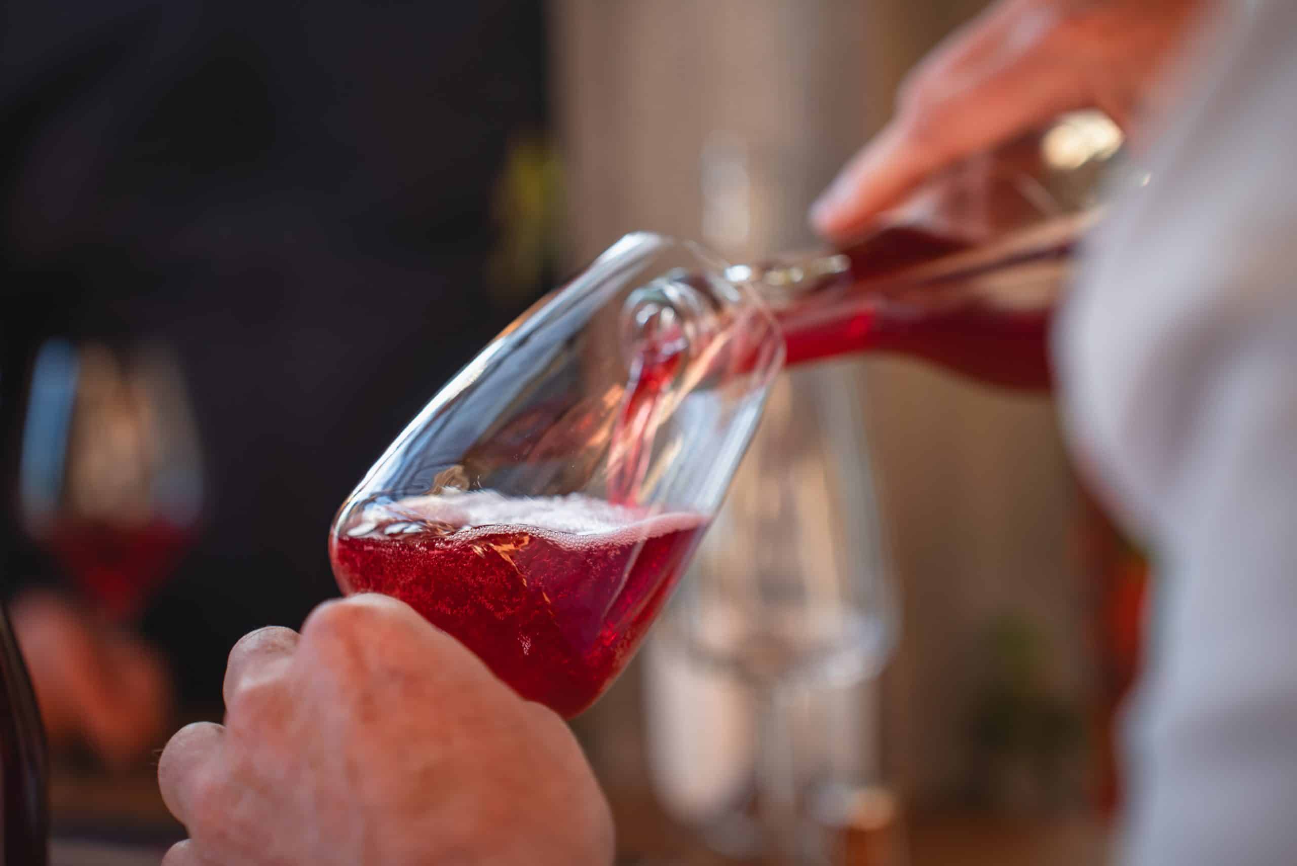 Un verre de vin rouge, tenu par une main, est rempli par une autre main versant le liquide pétillant d'une bouteille, dans un cadre intérieur flou.