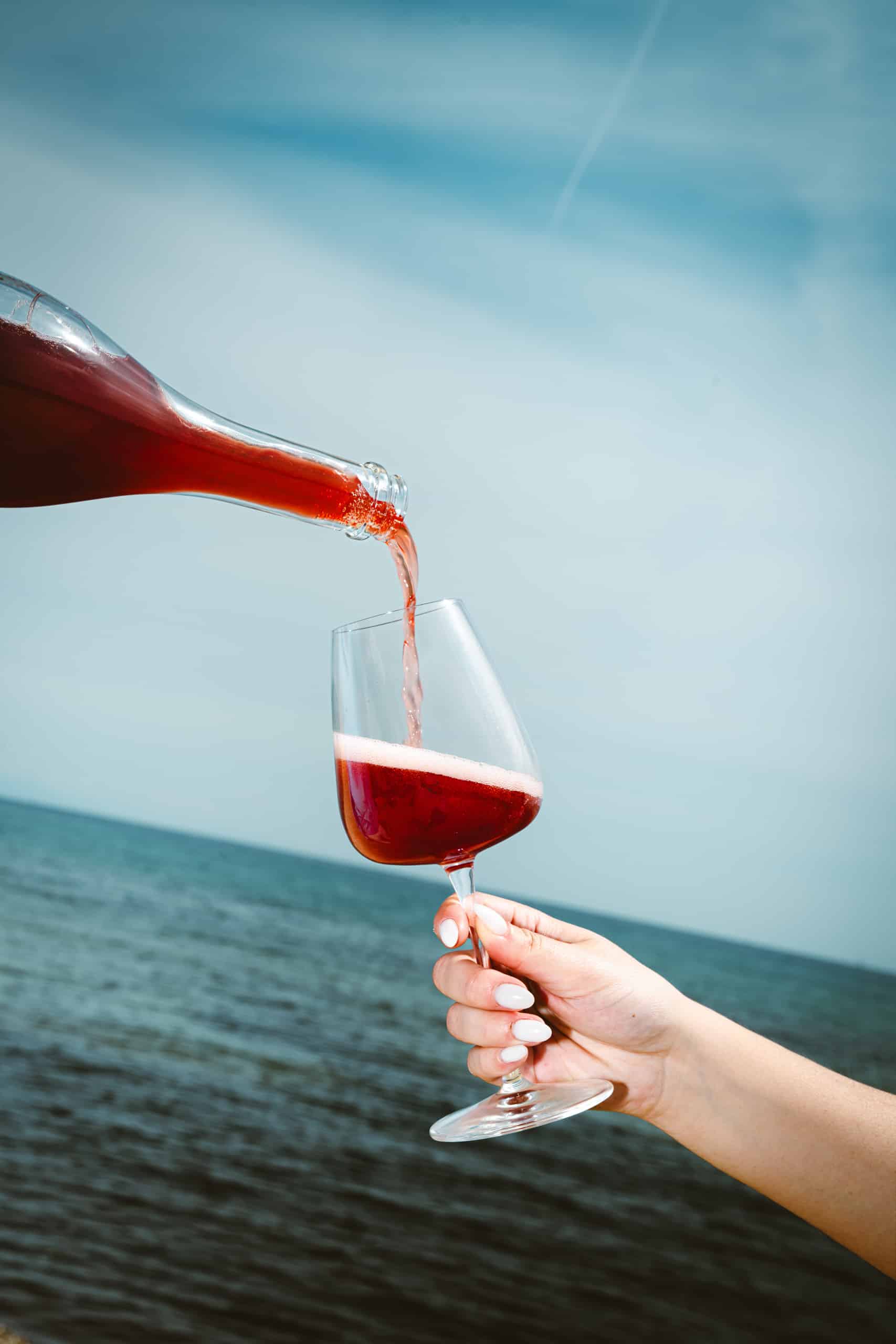 A hand holding a glass is being filled with red liquid poured from a bottle, with the sea and a clear blue sky in the background.