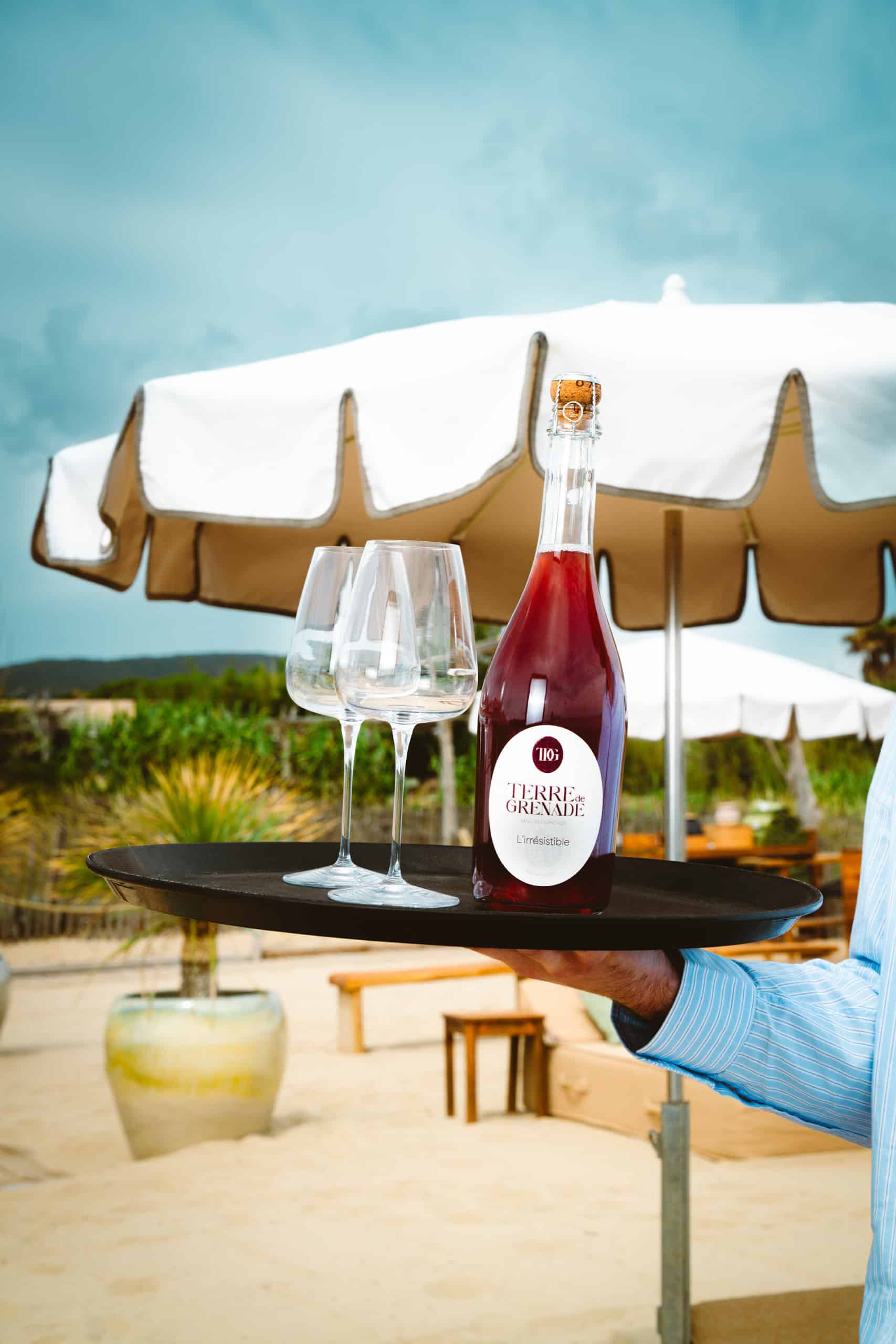 Un bras tient un plateau avec une bouteille de vin rouge pétillant et deux verres vides. Contexte extérieur sous un parasol blanc. Texte: "TERRE de GRENADE L'irrésistible".