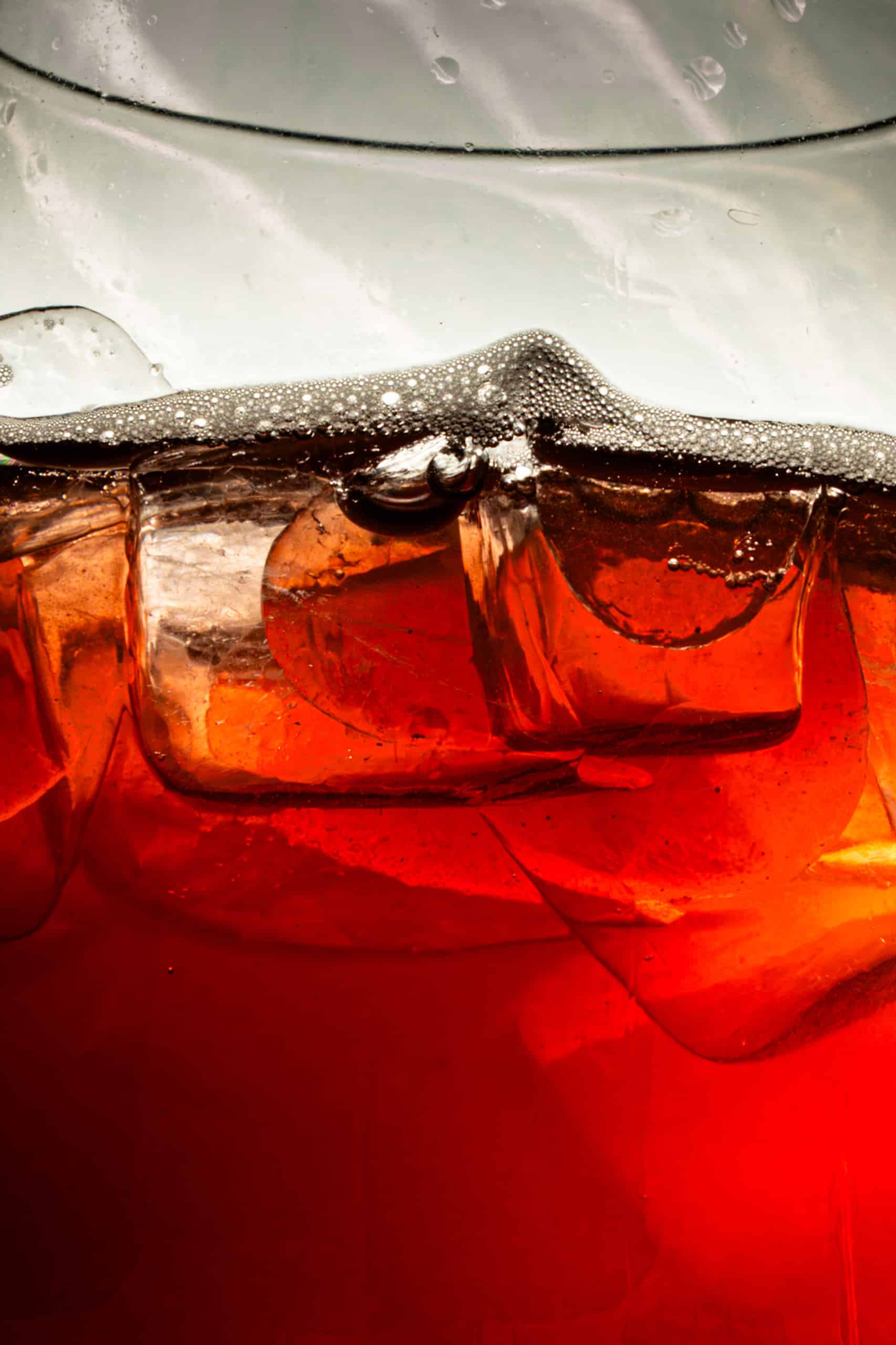 Ice cubes and foam floating in a dark red carbonated beverage, a close-up view inside a clear glass.
