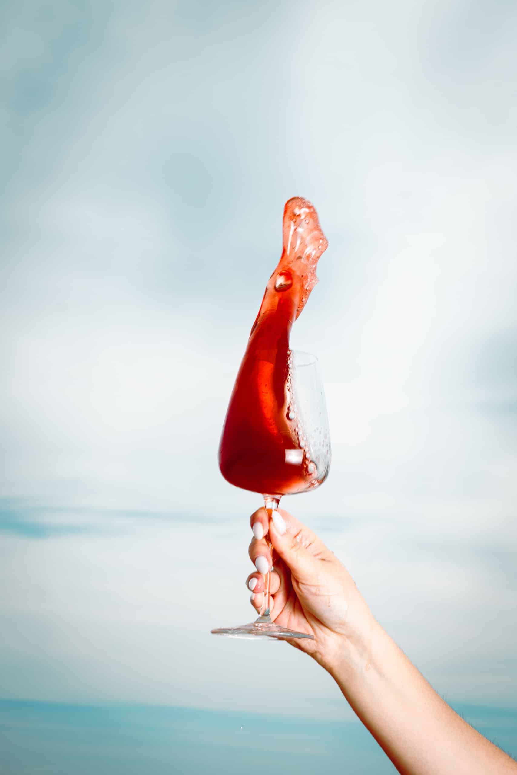 Main tenant un verre de vin rouge dont le liquide s'élève en une éclaboussure dynamique, sur fond bleu clair et flou.