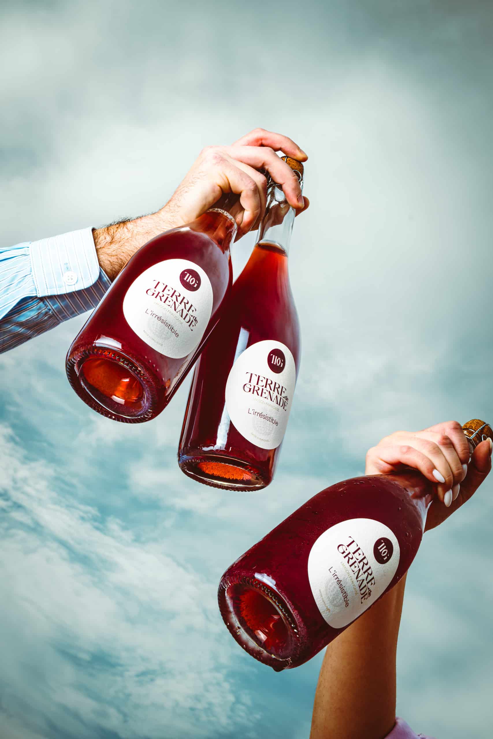 Trois bouteilles de boisson pétillante rosé sont tenues par trois mains sur un fond de ciel nuageux. Étiquettes: "TERRE de GRENADE", "MAISON FRANCAISE", "L'irrésistible".