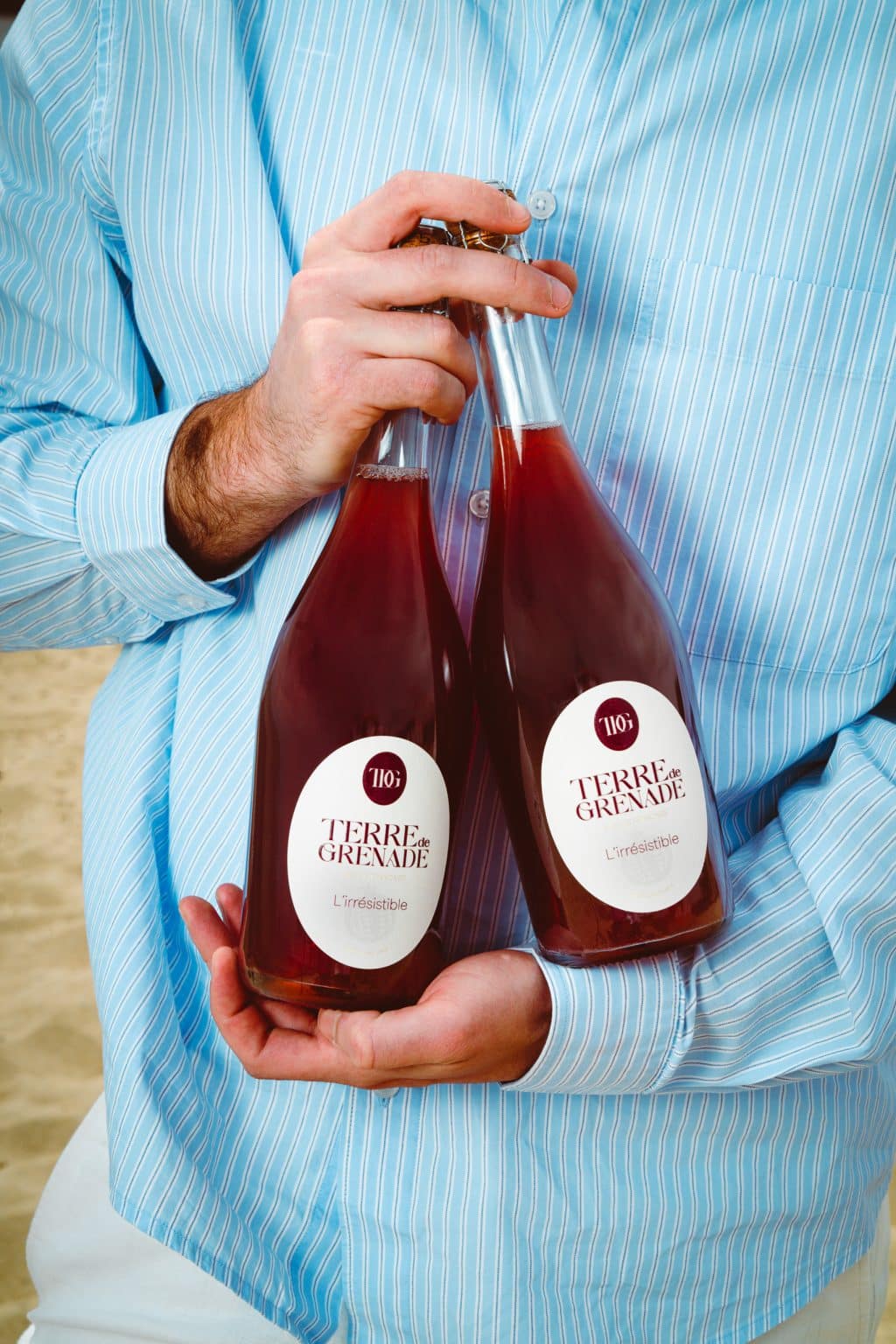 A man wearing a blue shirt with thin white stripes is holding two glass bottles filled with a red liquid. Their white labels read "TERRE de GRENADE L'irrésistible."