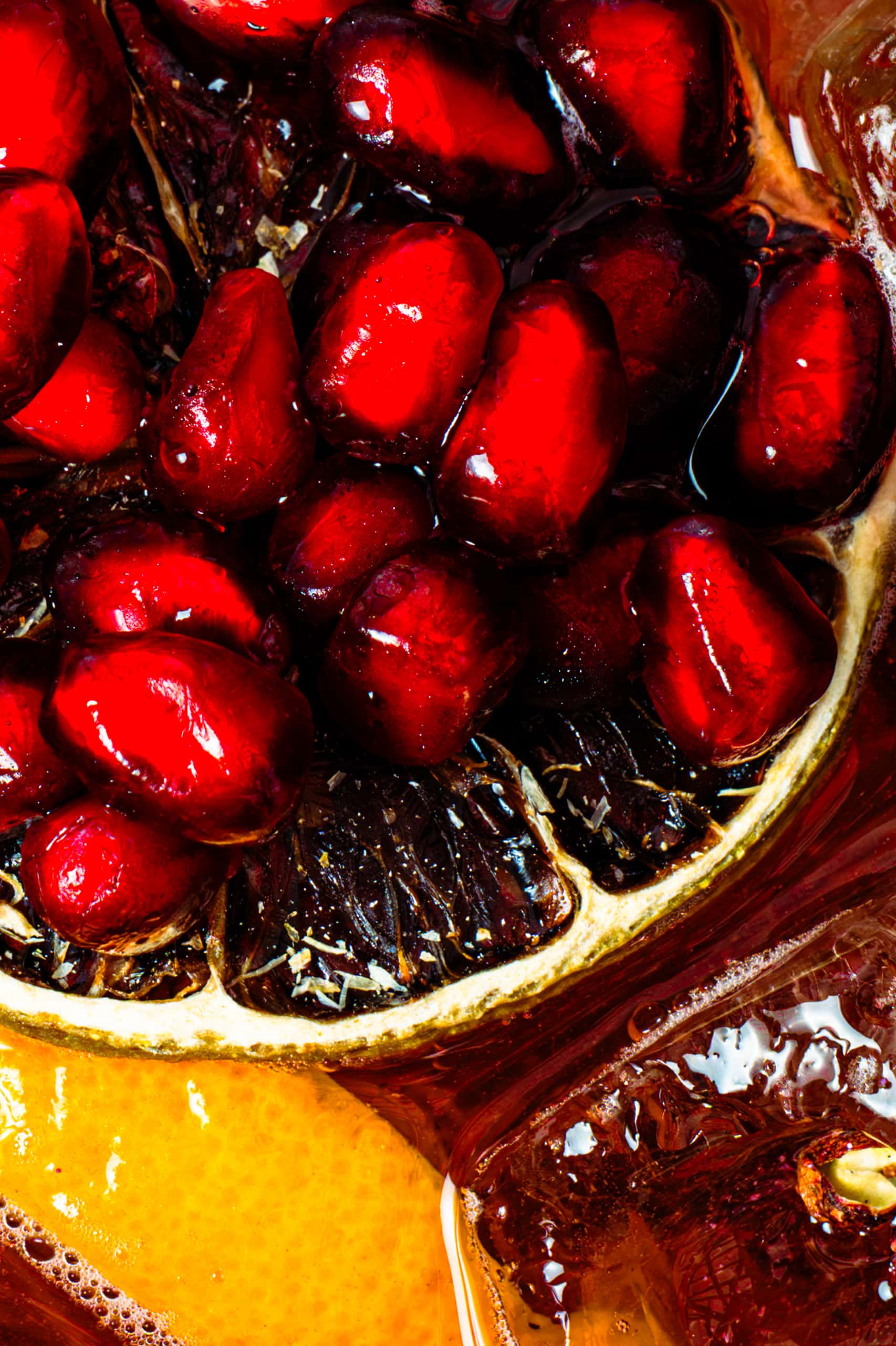 Arilles de grenade rouges, brillants et juteux, reposent sur une tranche de fruit sombre. Ils sont partiellement immergés dans un liquide rougeâtre, à côté d'une tranche d'agrume jaune.