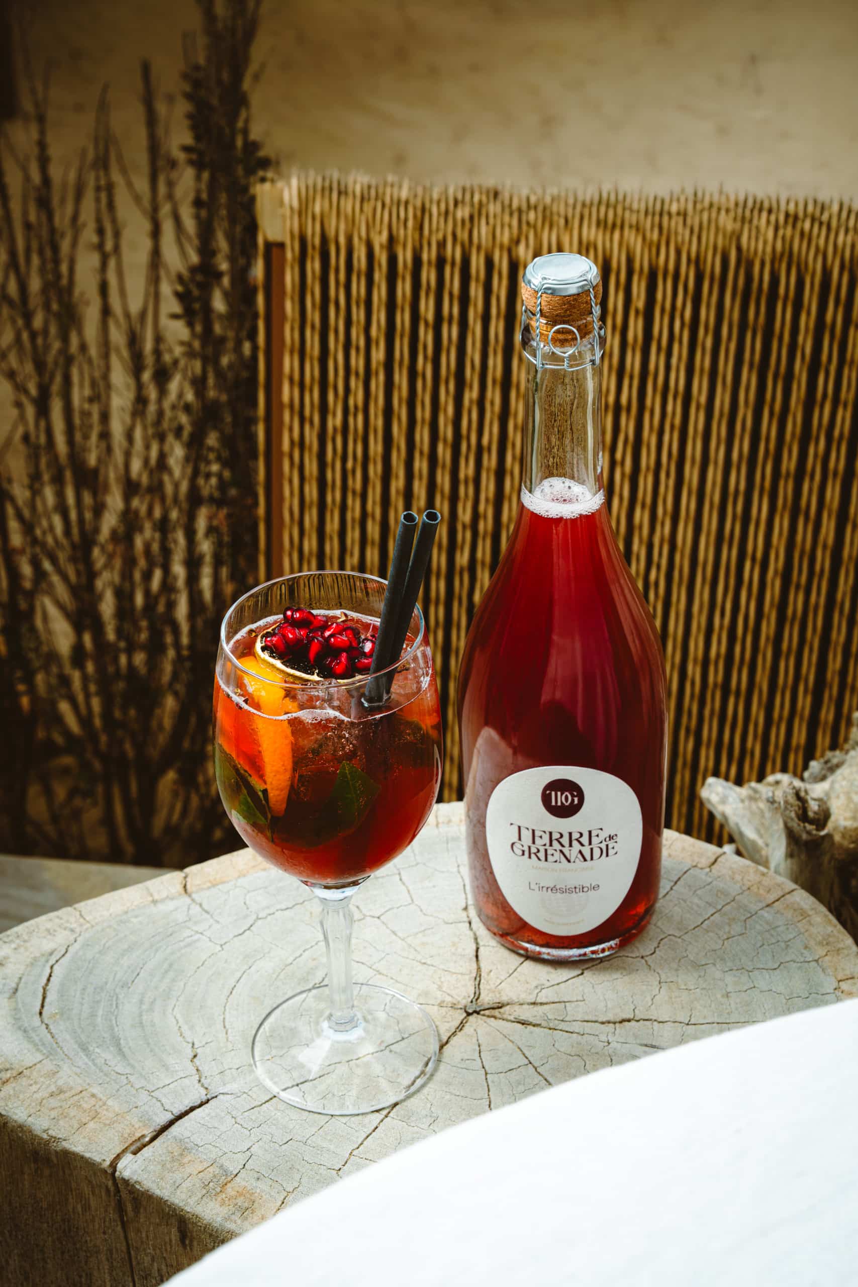 Red cocktail glass (pomegranate, orange, black straws) and a bottle of "TERRE de GRENADE MAISON FERRAND L'irrésistible." Placed on a wooden stump, natural background.