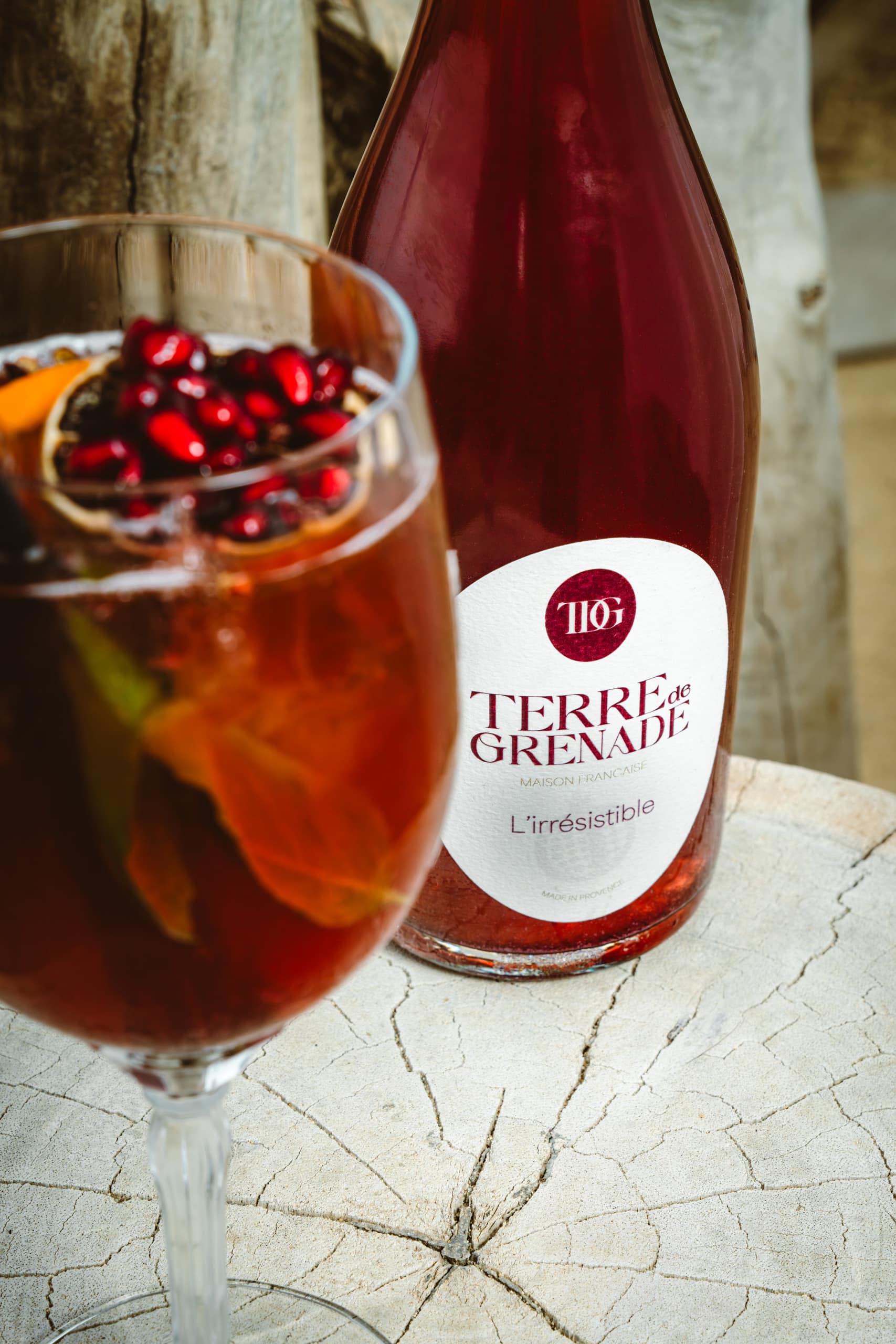 Un verre de boisson rouge garnie de grenade et d'orange, et une bouteille affichant "TERRE de GRENADE MAISON FRANÇAISE L'irrésistible MADE IN PROVENCE" reposent sur une surface claire et craquelée.