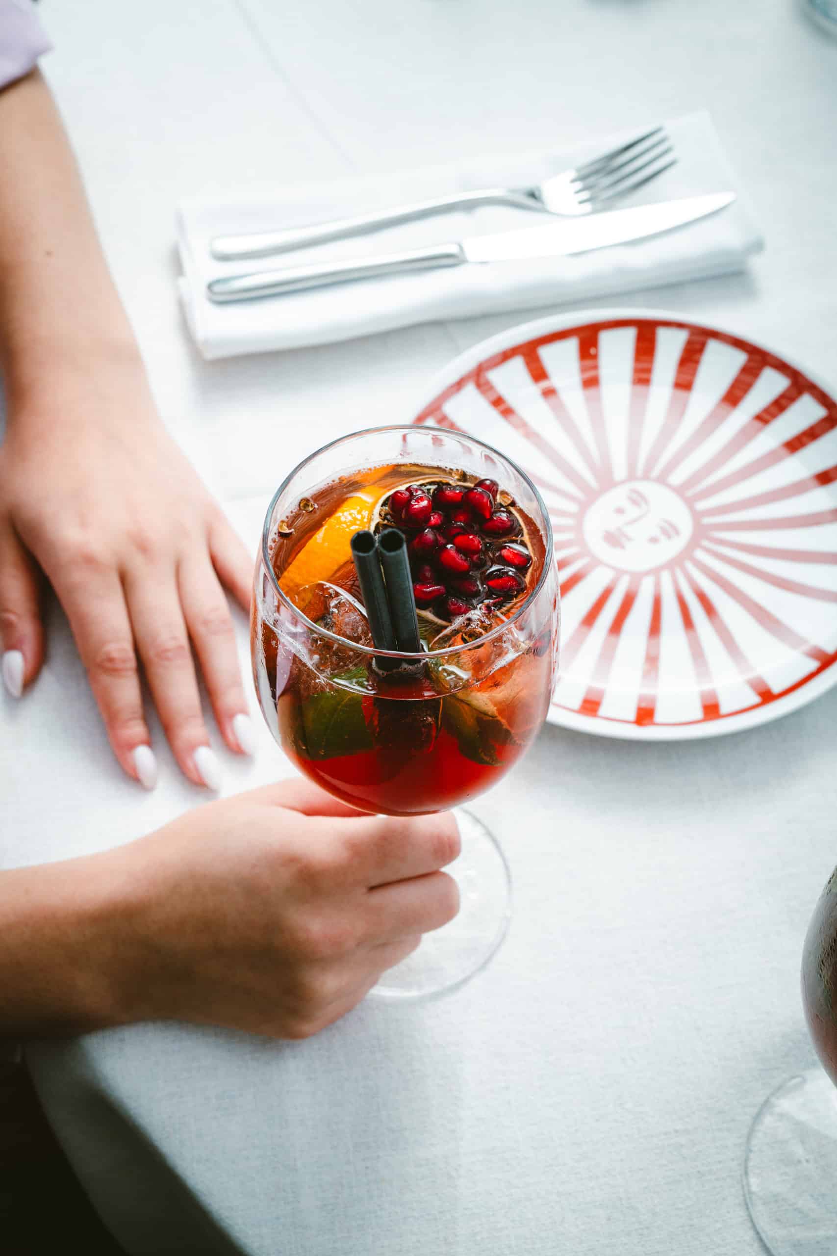 Verre de cocktail garni d'orange, menthe, grenade et deux pailles noires, tenu par une main. Sur une table blanche, à côté d'une assiette au motif soleil rouge et de couverts.