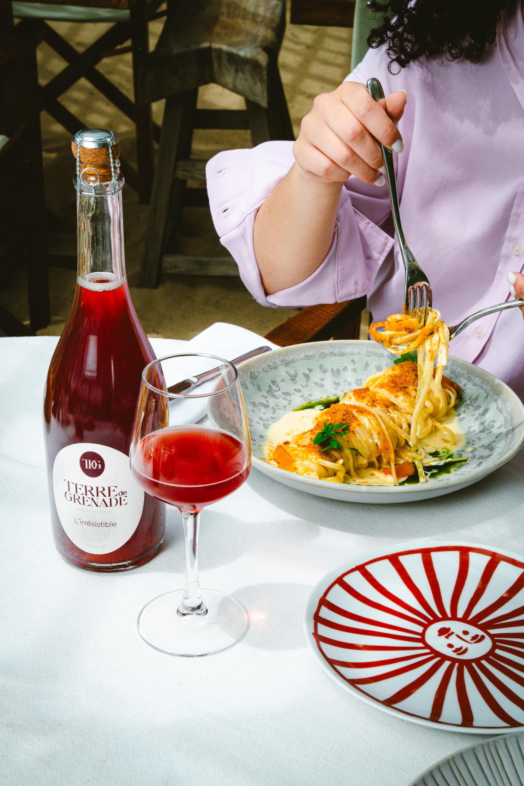 A woman's hand is eating spaghetti from a plate, next to a bottle of "TERRE de GRENADE MAISON FRANÇAISE L'irrésistible MADE IN FRANCE" and a glass of red drink, on a white tablecloth outdoors.