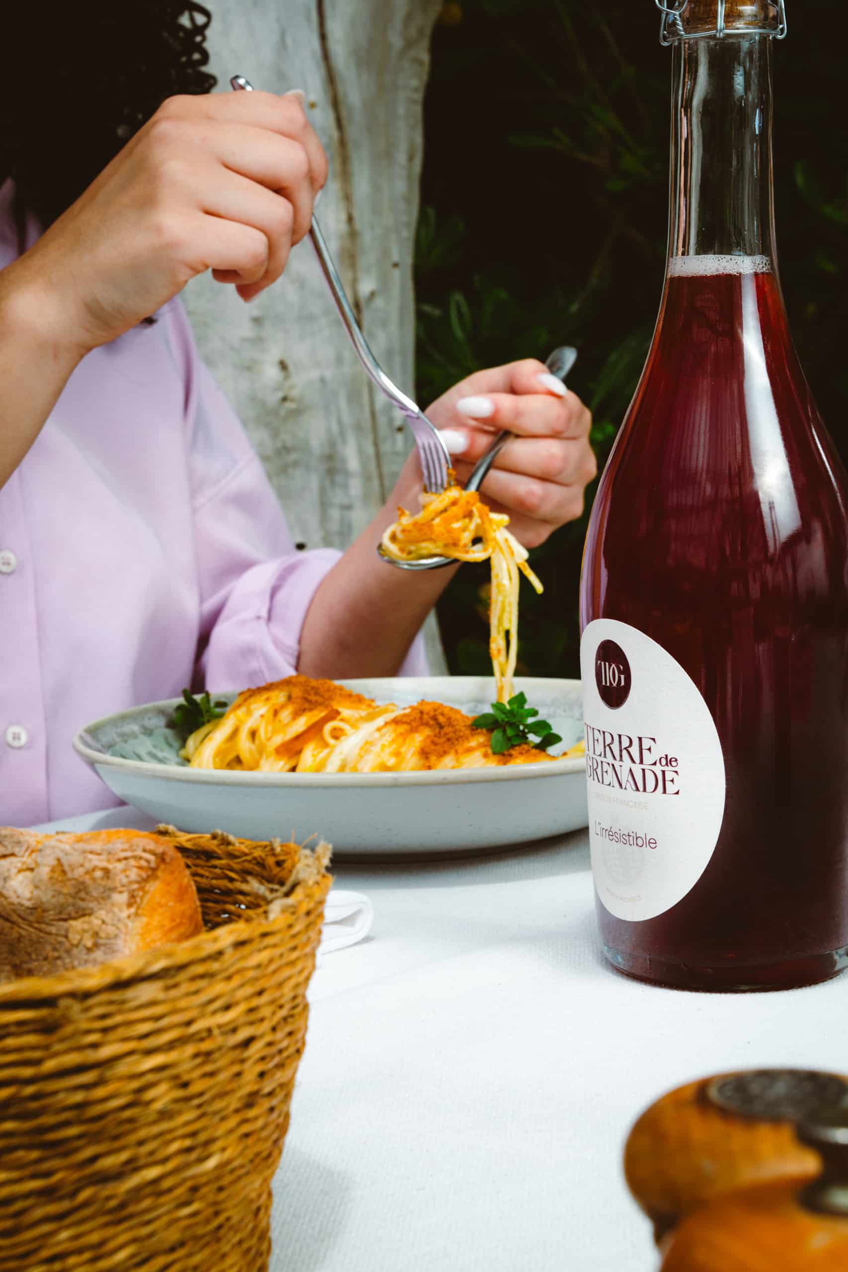 Mains féminines mangeant des pâtes d'une assiette sur une table, à côté d'une bouteille de "TERRE de GRENADE BOISSON FRANÇAISE L'irrésistible" et d'une corbeille de pain.