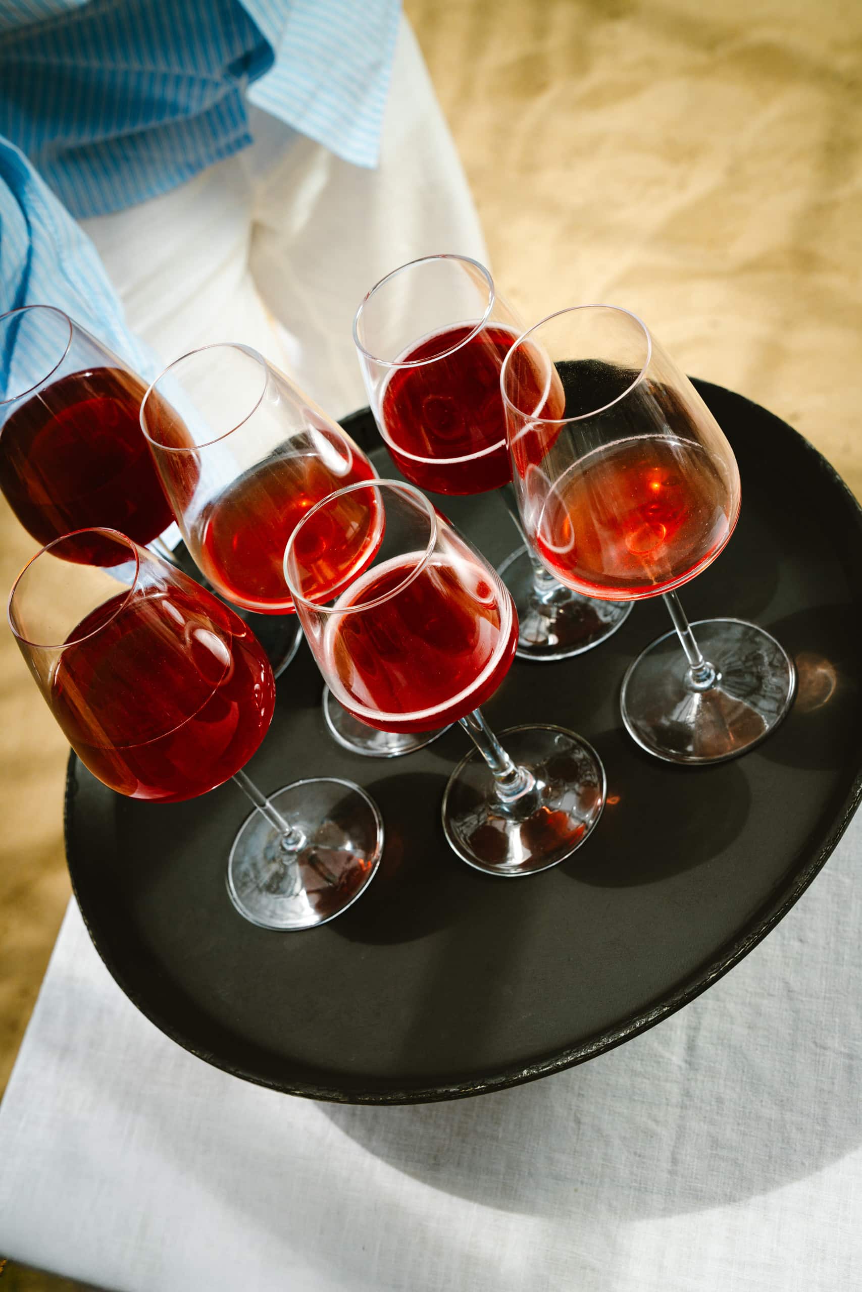 Six verres à pied de vin rosé sont servis sur un plateau rond noir, tenu par une personne vêtue d'une chemise rayée bleue.