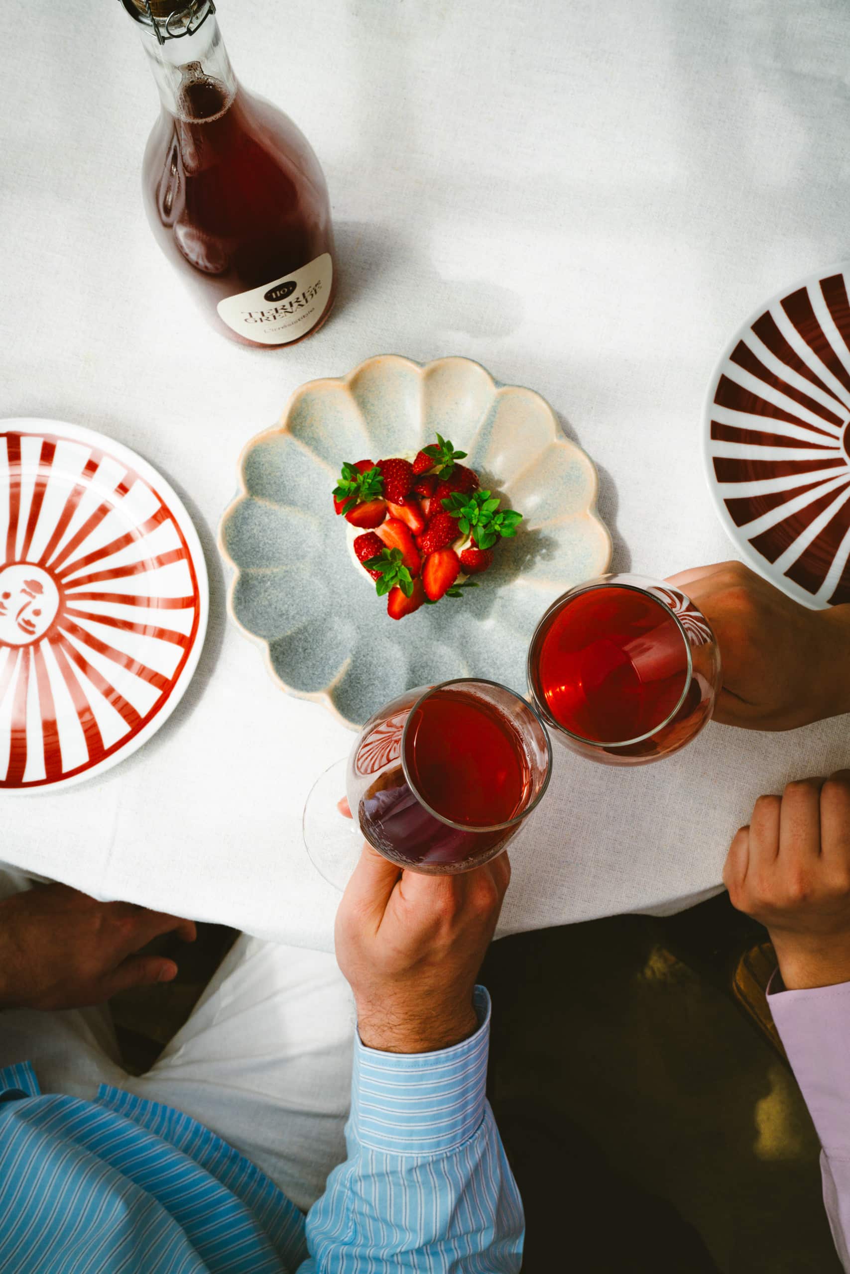 Two hands are clinking glasses filled with a red beverage. A bottle labeled “TERRE DE GRENADE L'irrésistible” and a plate of strawberries adorn the white table set with plates.