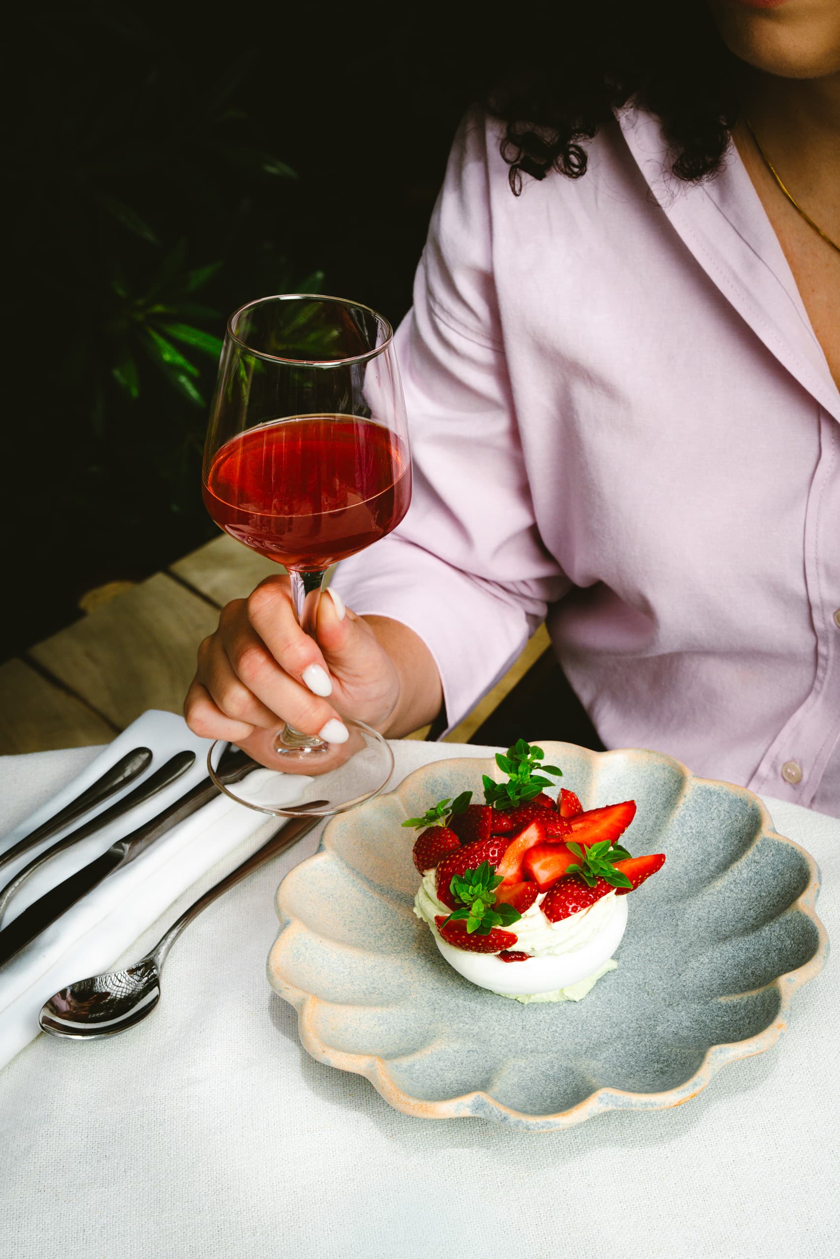 A hand holds a glass of sparkling pomegranate beverage next to a plate containing a strawberry and basil pavlova on a white table set with cutlery, against a dark, plant-filled background.