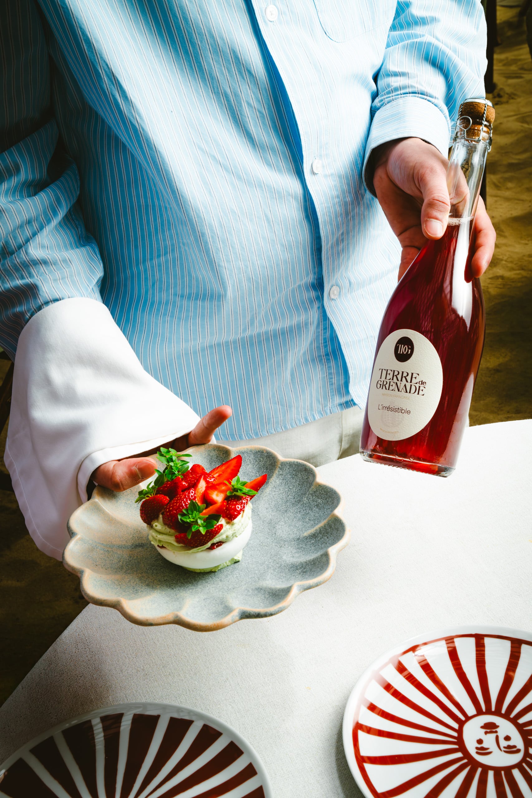Personne en chemise bleue présentant une coupe de fraises sur meringue et une bouteille de boisson rouge. Sur une table nappée avec assiettes décorées. La bouteille indique "TERRE de GRENADE MAISON FRANCAISE L'irrésistible".