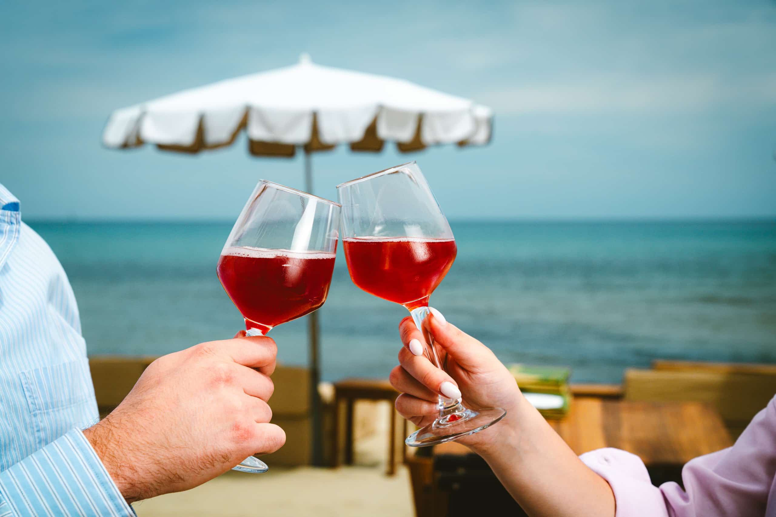 Two hands clink glasses of sparkling pomegranate beverage. The scene takes place by the sea, under a white beach umbrella.