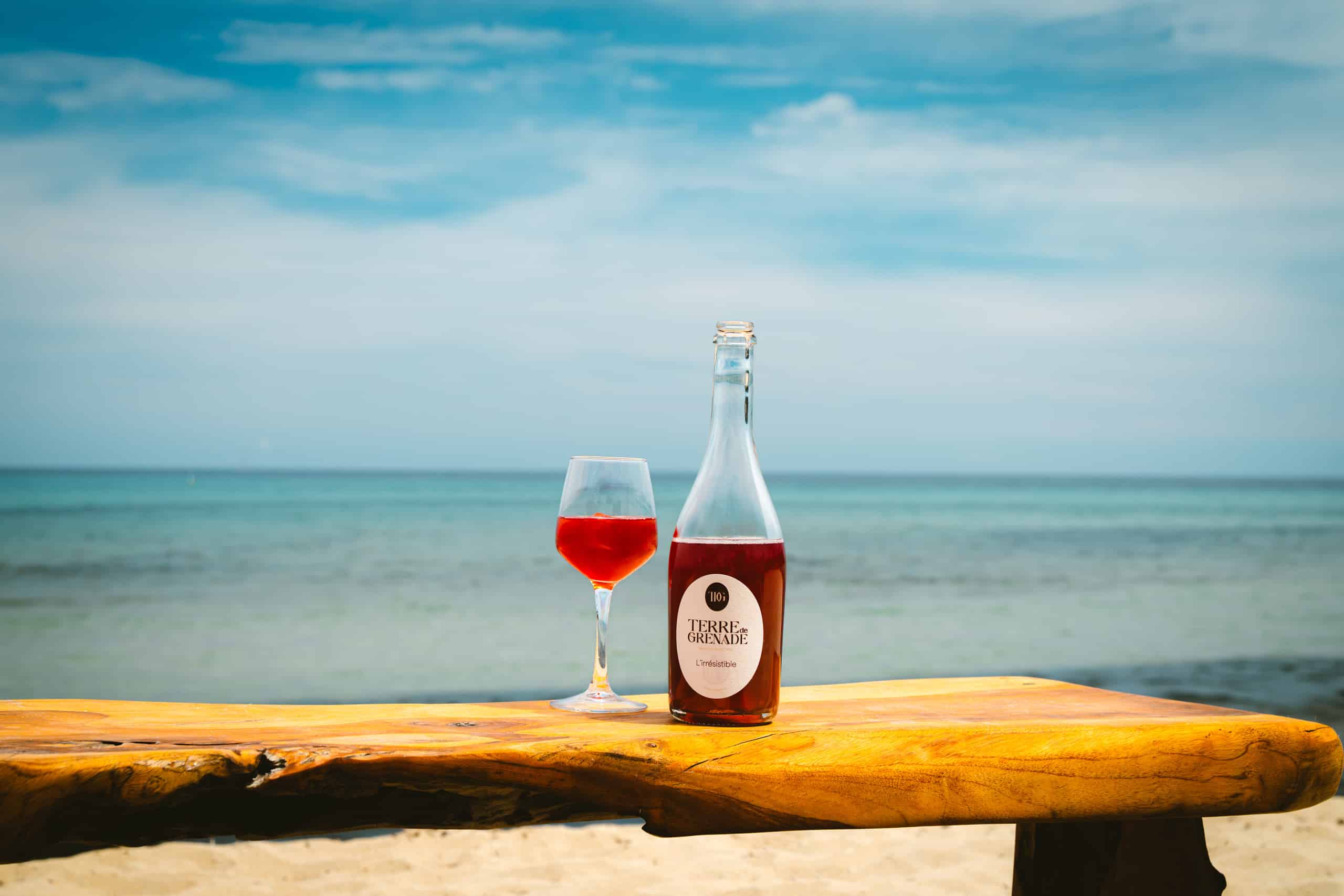 Un verre de boisson rouge et une bouteille, étiquetée "TERRE de GRENADE L'irrésistible", sont posés sur une table en bois sur une plage, face à la mer et au ciel.
