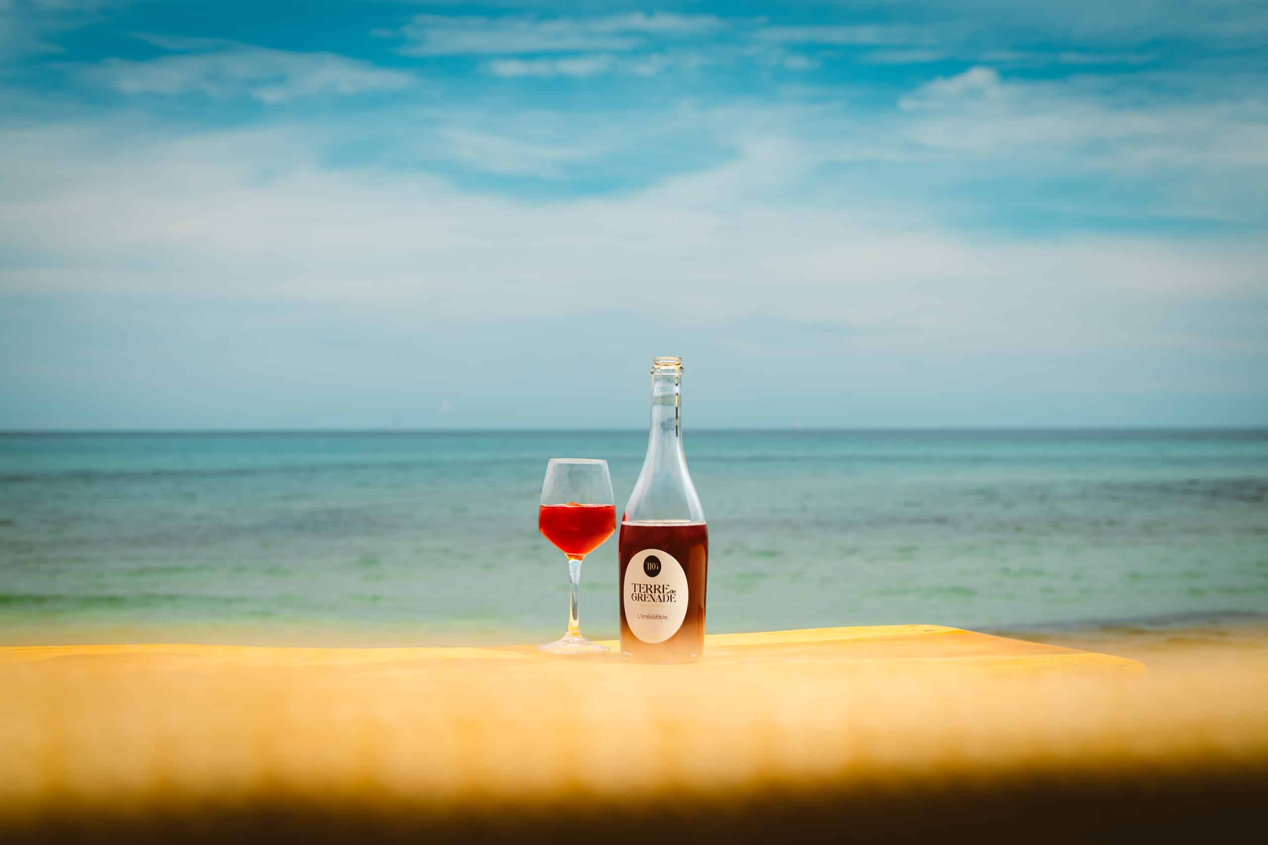 A glass of red liquid and a "TERRE de GRENADE" ("L'irrésistible") bottle sitting on a light-colored surface, facing a blue ocean under a cloudy sky.