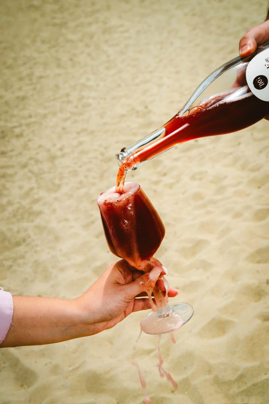 A sparkling red liquid is poured from a labeled bottle into a glass held in a hand. The liquid overflows, spilling onto the hand and the surrounding sand.