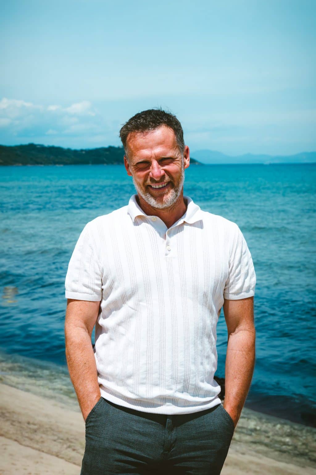 A middle-aged, bearded man, smiling with his eyes closed, his hands in his pockets. He is standing on a rocky beach, facing the blue sea and a distant mountainous horizon.