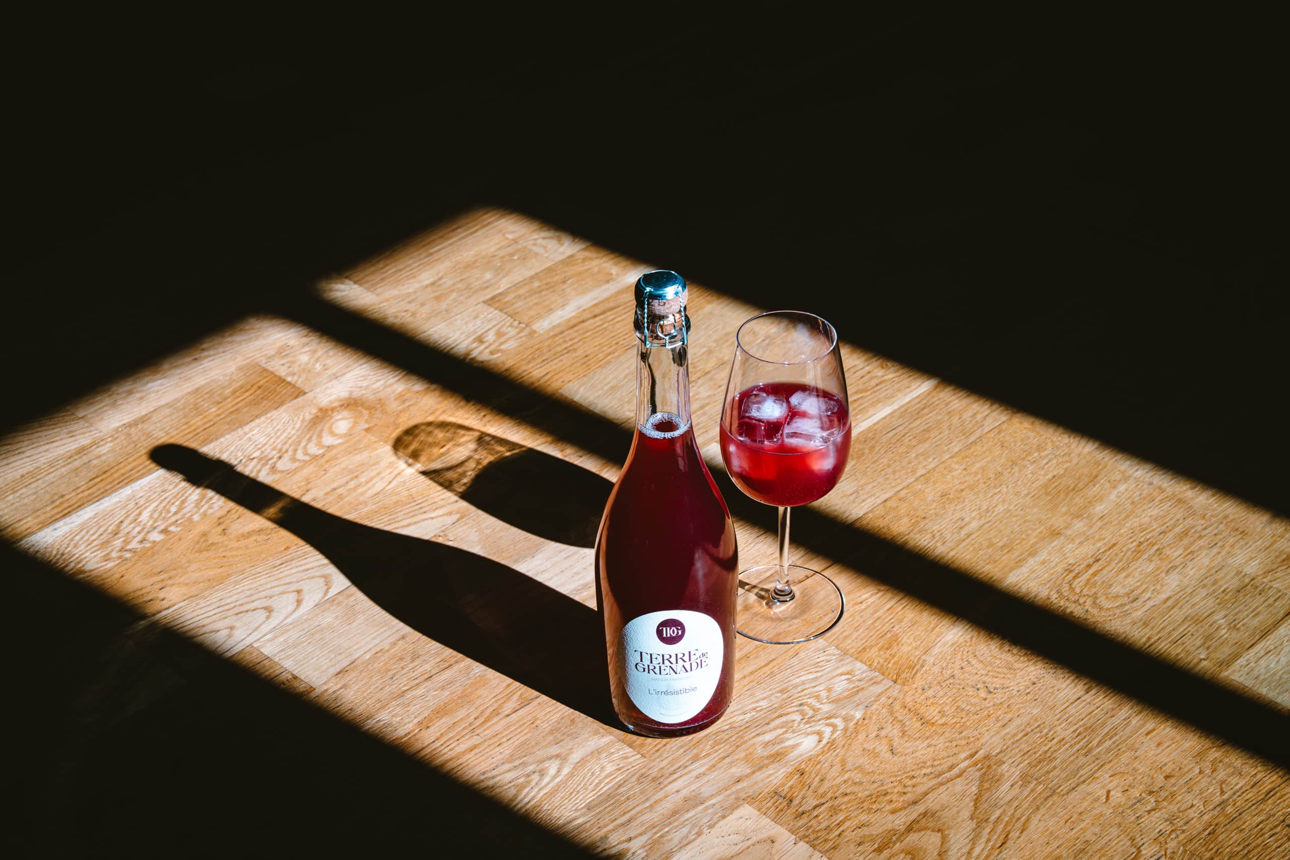 Bouteille de boisson rouge pétillante et verre avec liquide et glaçons, sur parquet en bois clair. Un rayon de soleil diagonal éclaire partiellement la scène, projetant des ombres nettes. Texte: "TERRE de GRENADE MAISON ROUGE L'irrésistible".