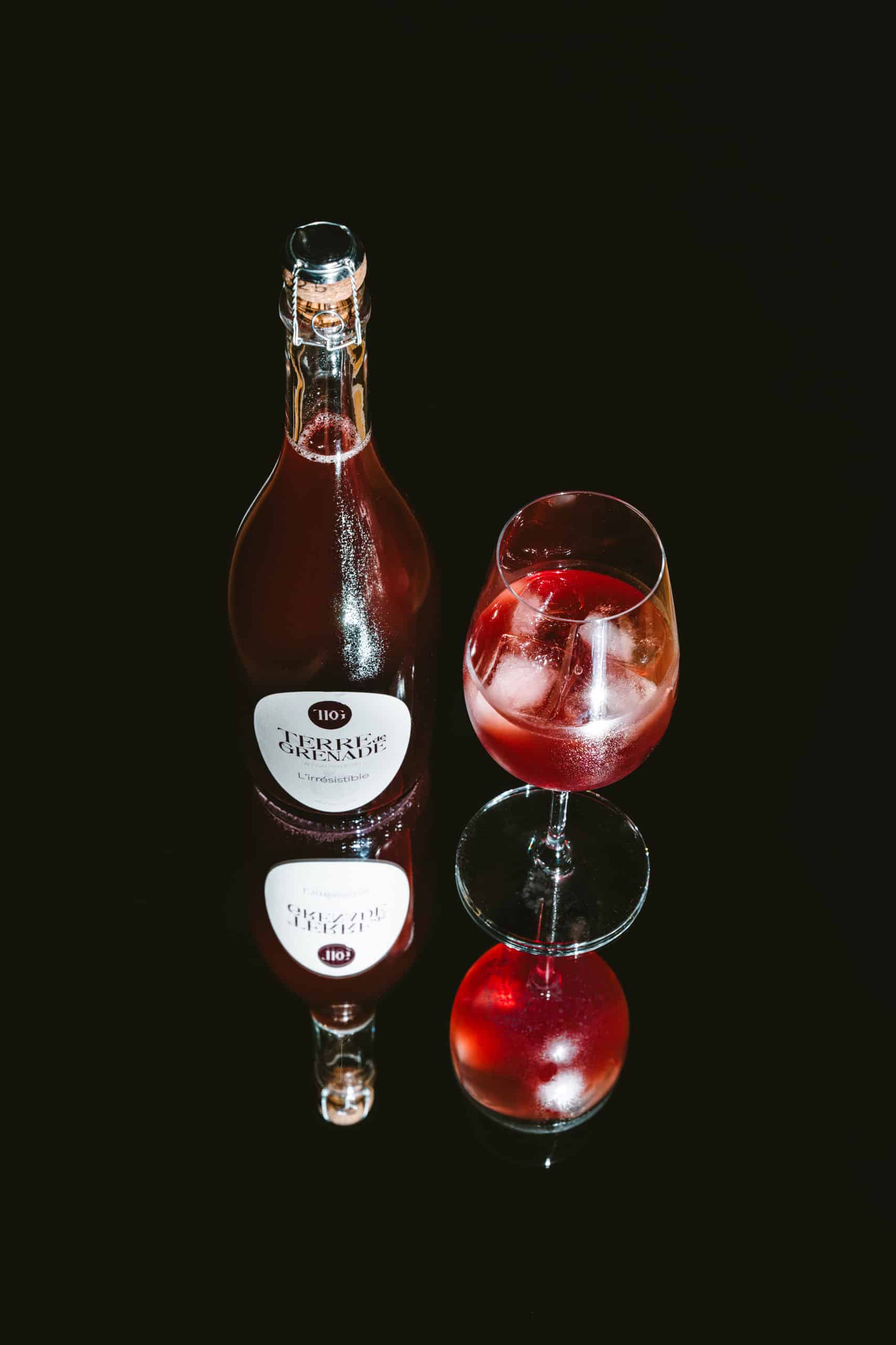 Une bouteille de boisson rouge pétillante est placée à côté d'un verre rempli de la même boisson avec des glaçons, sur une surface noire réfléchissante. L'étiquette de la bouteille indique : "TERRE de GRENADE L'irrésistible".