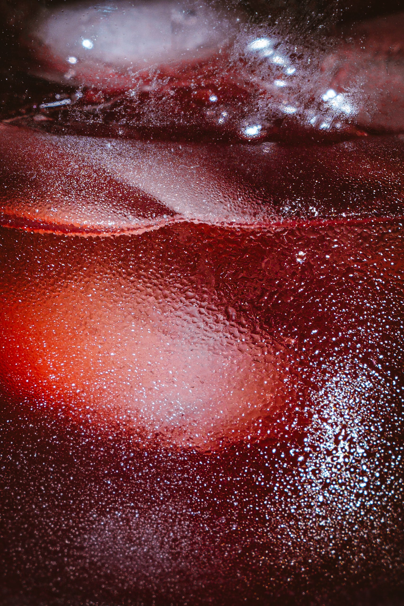 A glass of pomegranate sparkling beverage with ice cubes. Its surface is covered with condensation and bubbles, with intense, shimmering reflections.