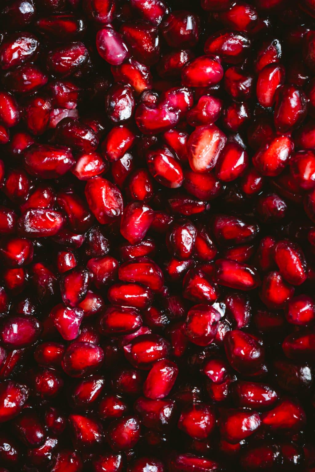 Bright red pomegranate seeds, clustered together, fill the entire frame against a dark background.
