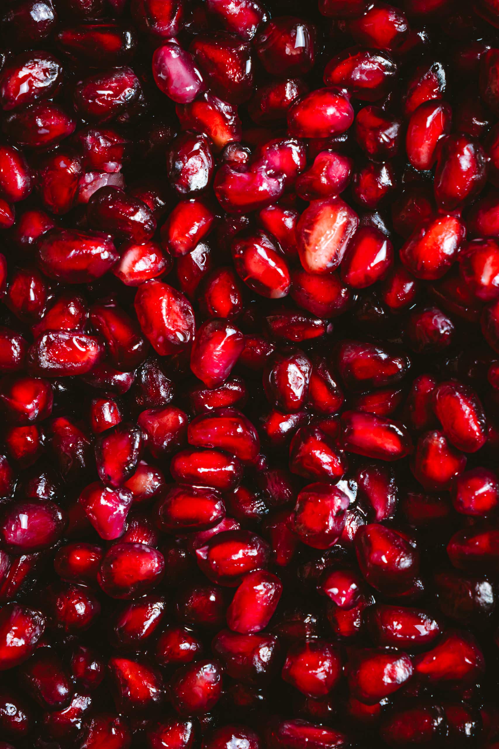 Bright red pomegranate seeds, clustered together, fill the entire frame against a dark background.